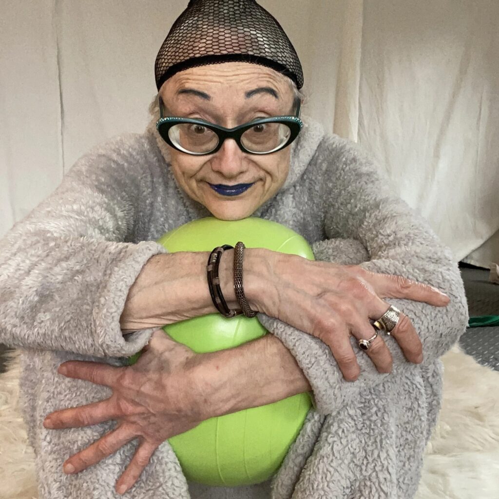 Character in fuzzy grey onesie with bold green glasses and blue lips hold green ball.